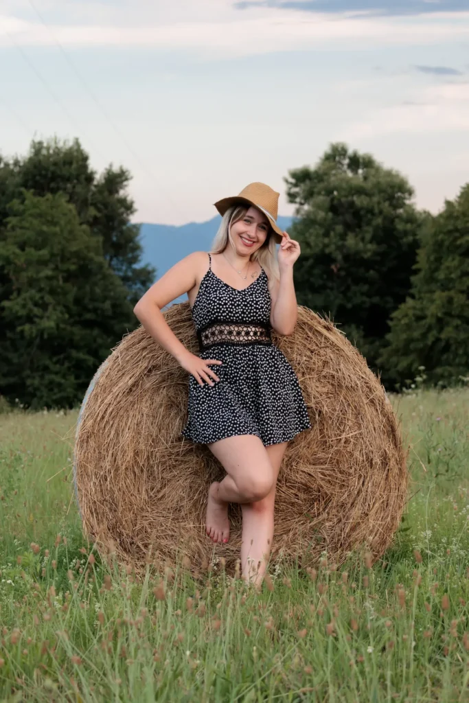 Une jeune fille posant avec un chapeau dans un champ, appuyée contre une botte de foin.